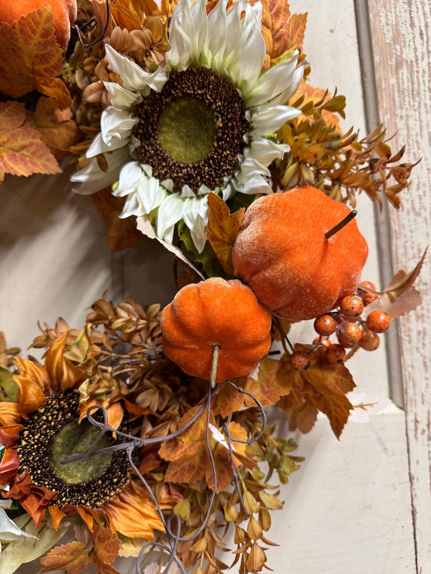 Autumn floral wreath with faux sunflowers, orange velvet pumpkins, fall leaves, and berry accents on a white distressed wood background.