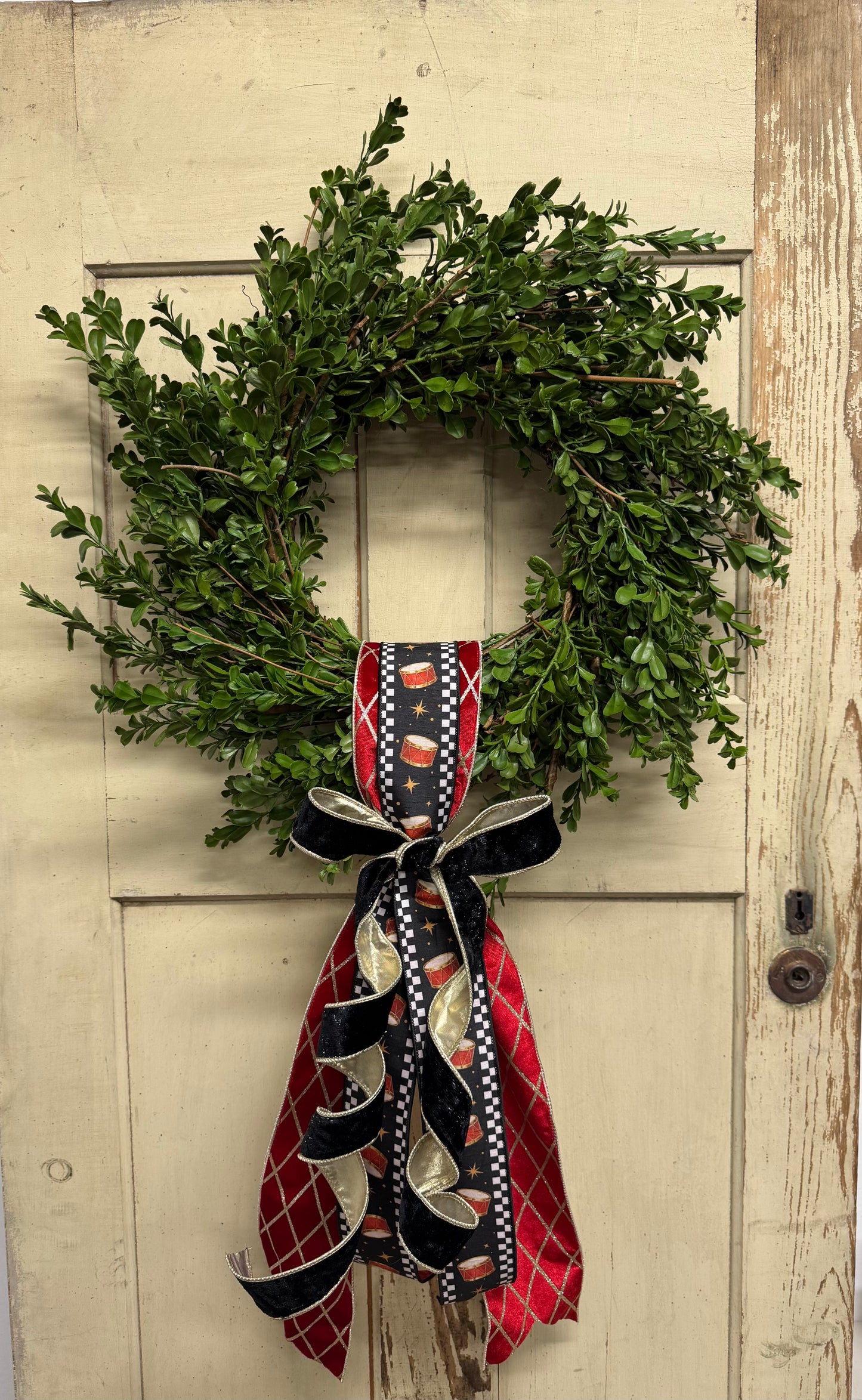  green boxwood Christmas wreath with red drum-print ribbon, gold accents, and a black velvet bow with cascading tails.”