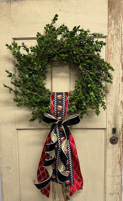  green boxwood Christmas wreath with red drum-print ribbon, gold accents, and a black velvet bow with cascading tails.”