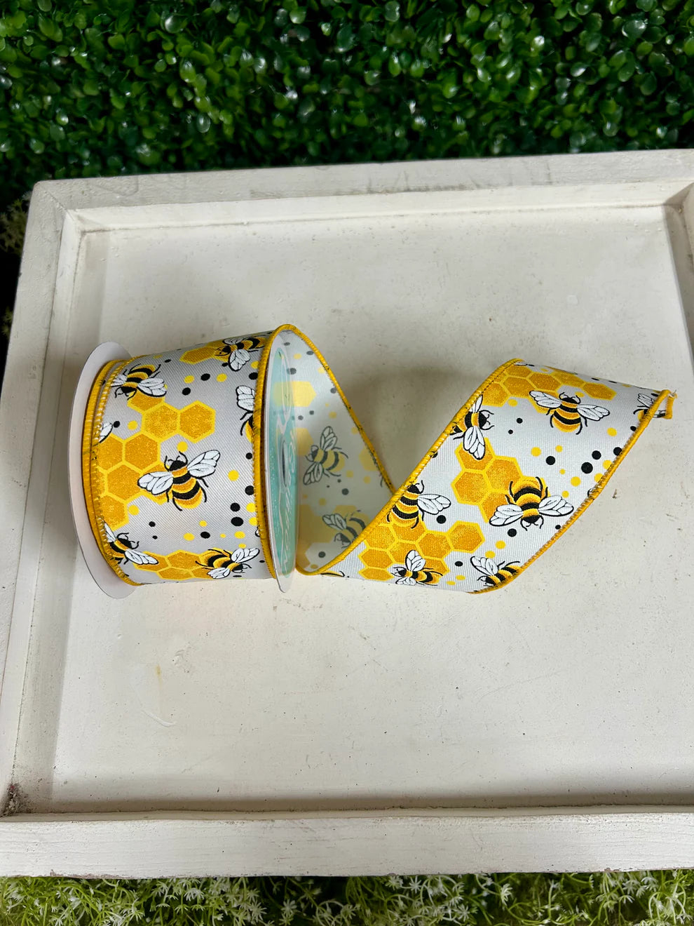 2.5 inch wired white ribbon with bumblebee and honeycomb print in yellow and black, rolled on a white decorative tray with green boxwood backdrop