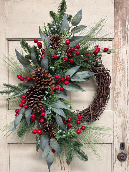 Christmas grapevine wreath with green pine and eucalyptus, red and blue berries, and brown pinecones.”