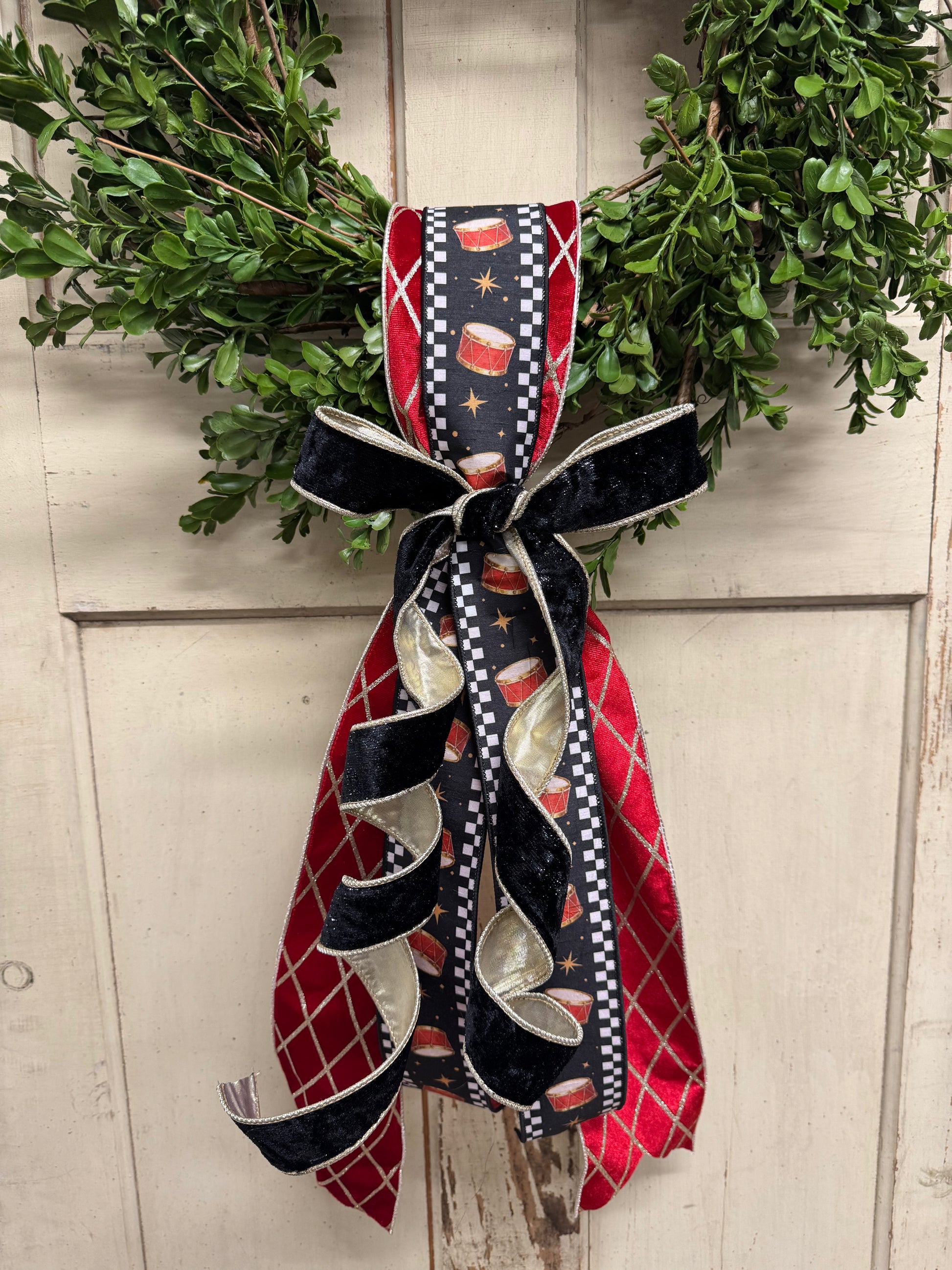 green boxwood Christmas wreath with red drum-print ribbon, gold accents, and a black velvet bow with cascading tails.”