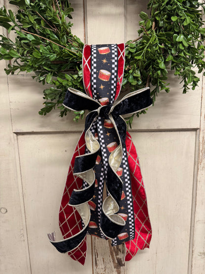 green boxwood Christmas wreath with red drum-print ribbon, gold accents, and a black velvet bow with cascading tails.”