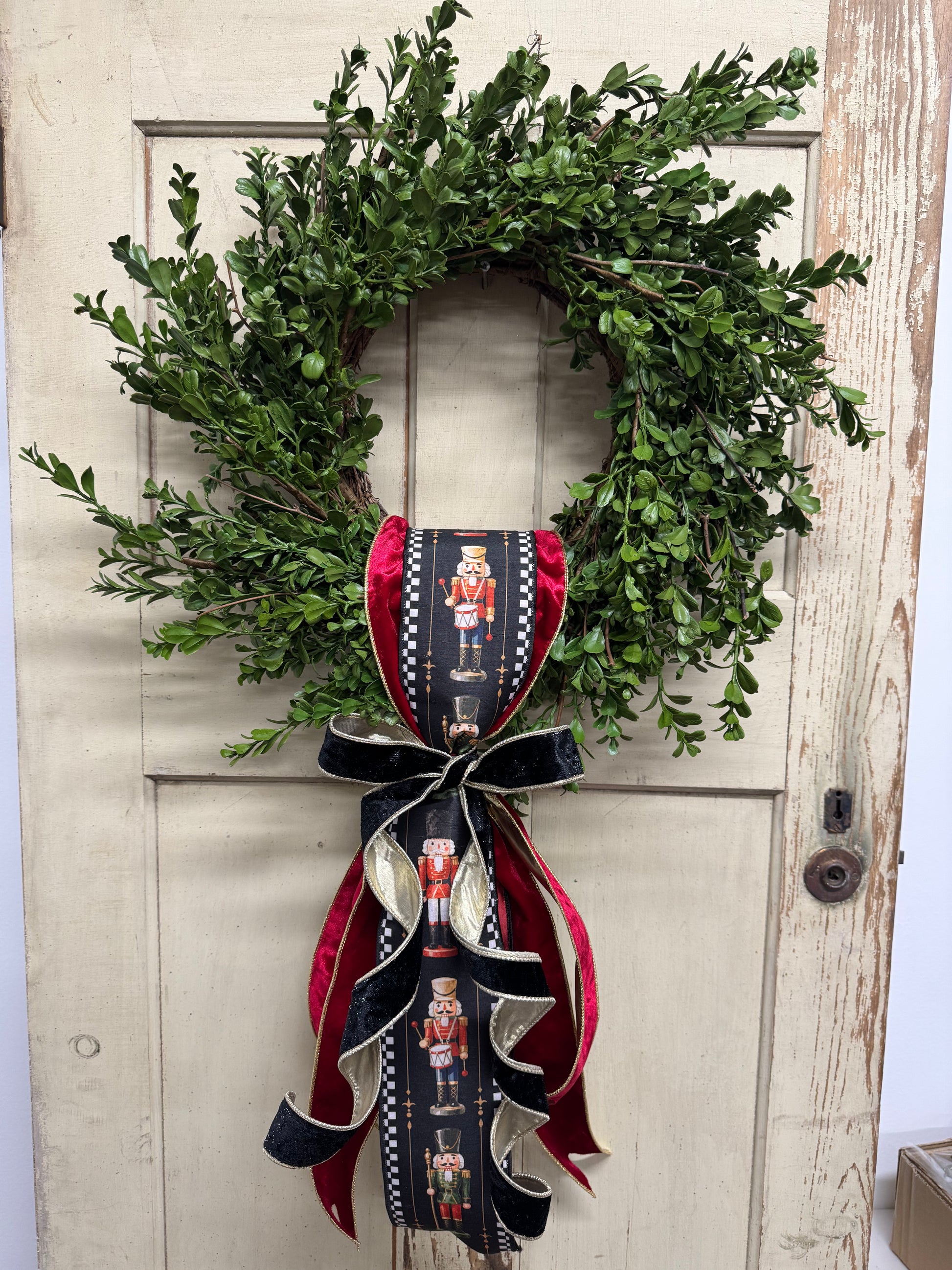 Green boxwood grapevine Christmas wreath with a nutcracker-print ribbon sash, red velvet ribbon, and a gold-edged black velvet bow.”