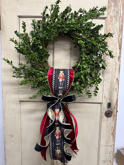Green boxwood grapevine Christmas wreath with a nutcracker-print ribbon sash, red velvet ribbon, and a gold-edged black velvet bow.”