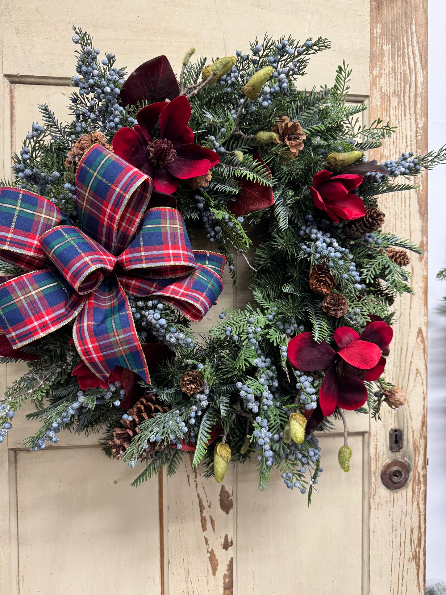 Christmas pine wreath with red magnolias, burgundy leaves, blue juniper berries, pinecones, and a red, navy, and green plaid bow.”