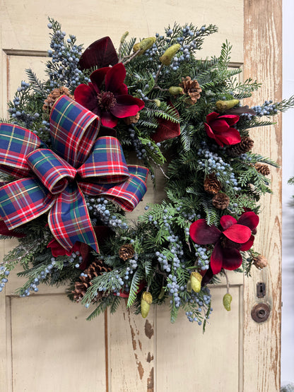 Christmas pine wreath with red magnolias, burgundy leaves, blue juniper berries, pinecones, and a red, navy, and green plaid bow.”