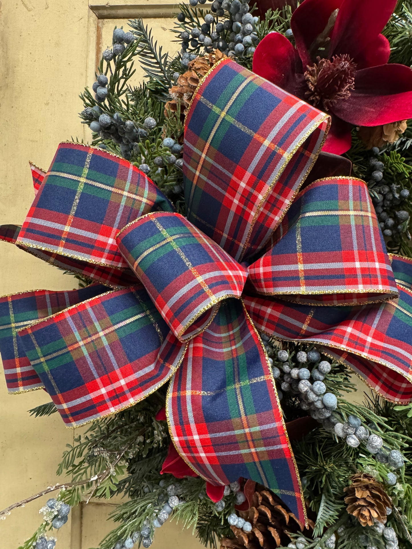 Red, navy blue, and green tartan plaid bow with gold detailing, surrounded by evergreen foliage, blue berries, and red magnolias.”