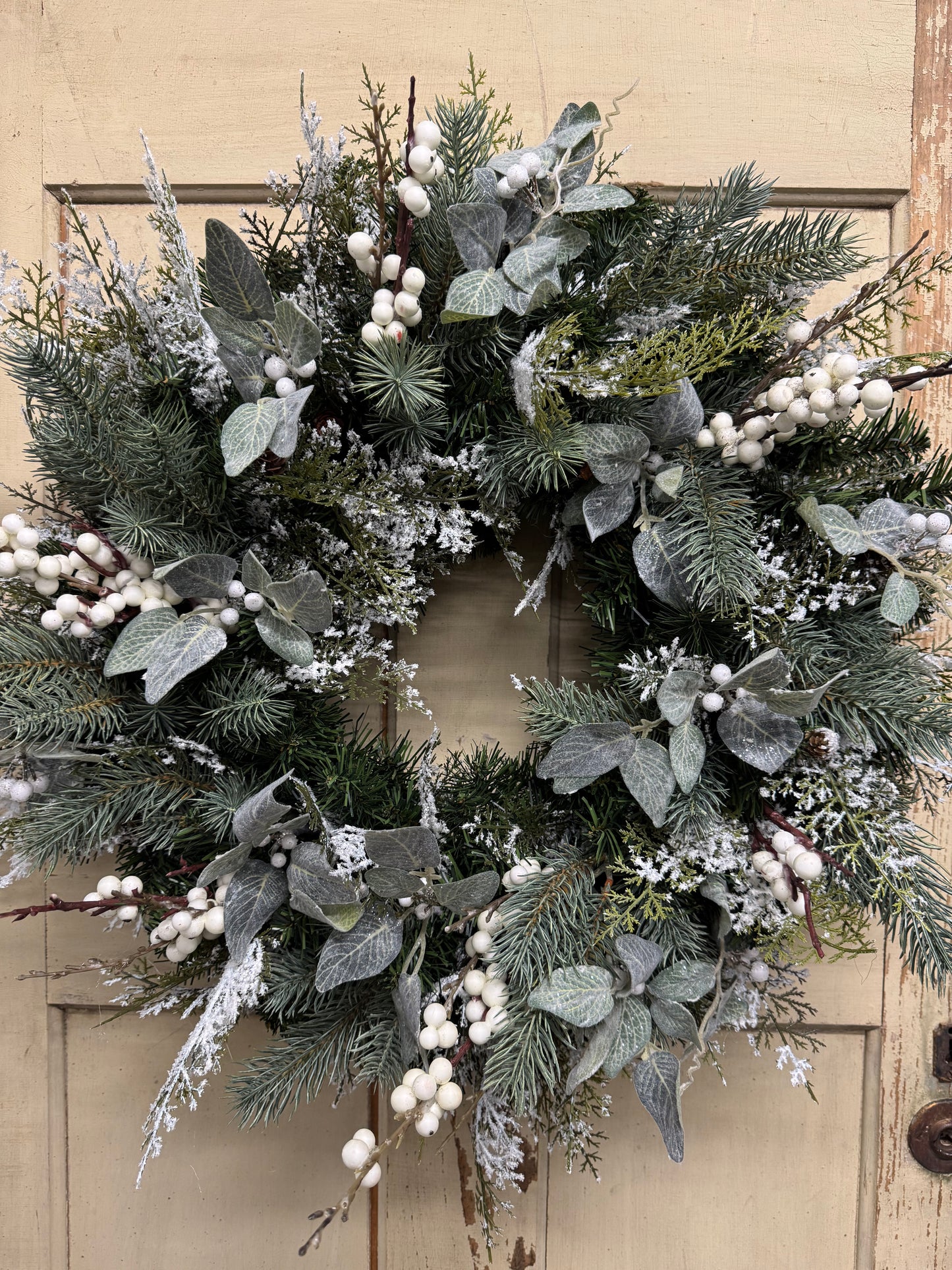 23-inch snowy winter wreath with green faux pine, snowy cedar, muted green leaves, and white berries.”