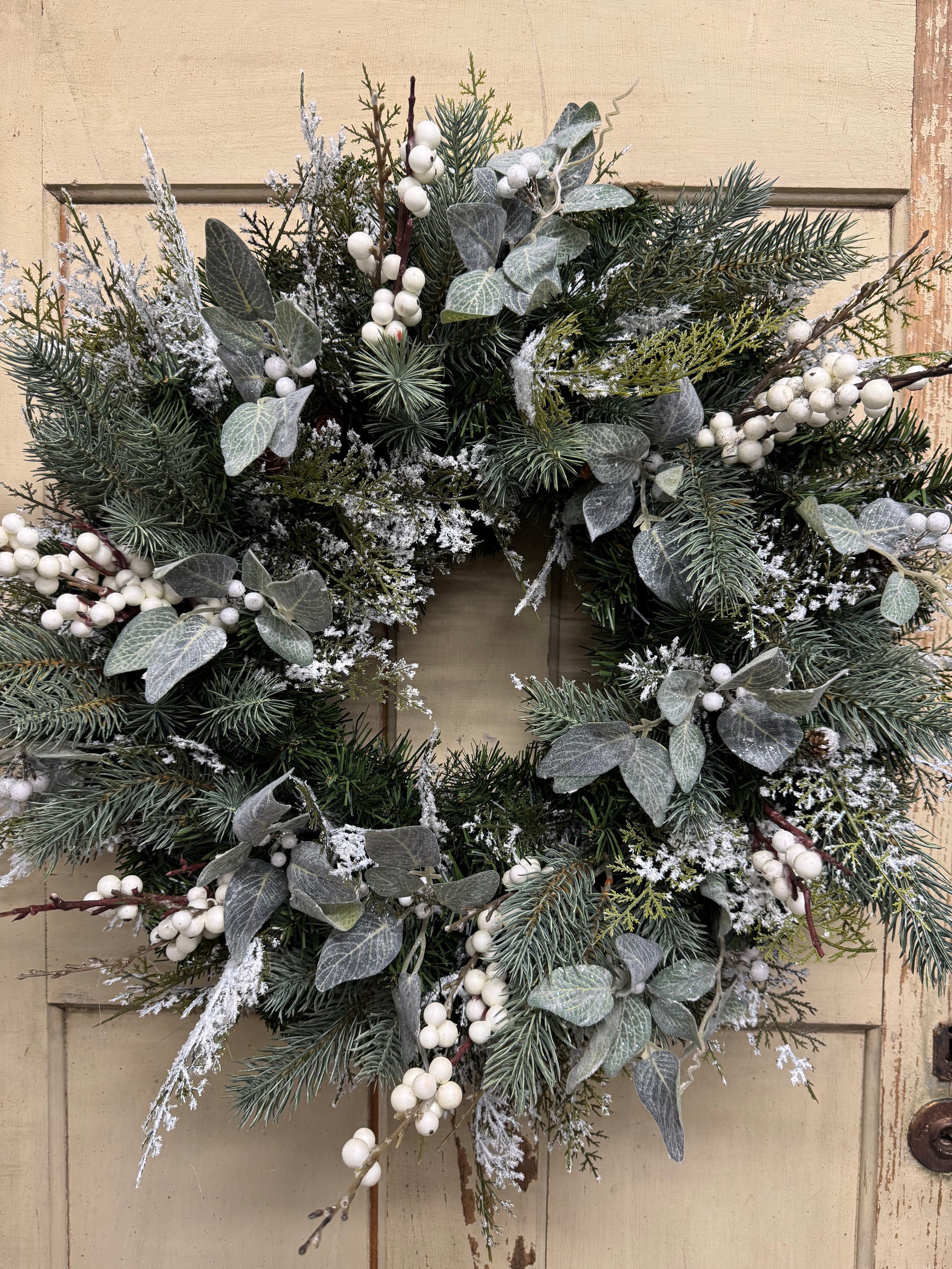 23-inch snowy winter wreath with green faux pine, snowy cedar, muted green leaves, and white berries.”