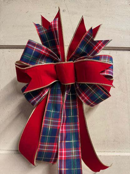 A large Christmas wreath bow made from red ribbon and red, blue, green, and yellow plaid ribbon, with gold trim, attached to a light-colored wooden door.
