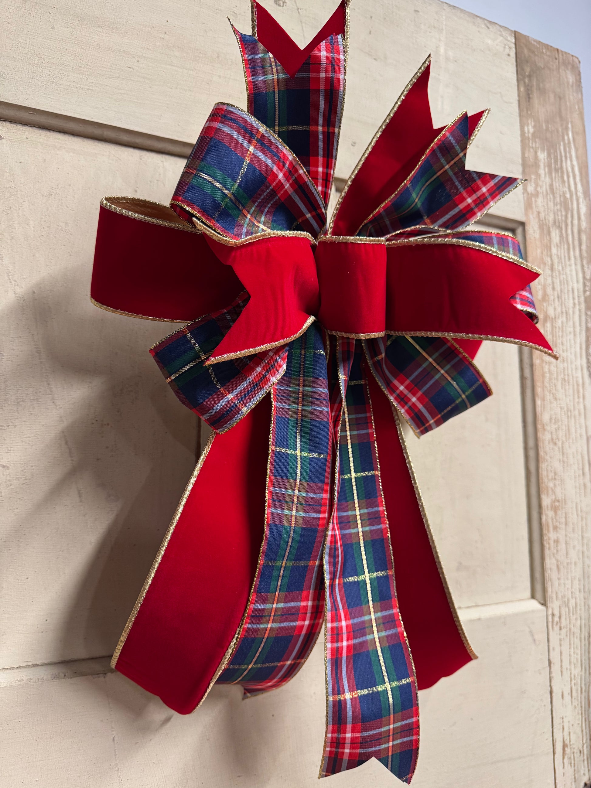 A large Christmas wreath bow made from red velvet ribbon and red, blue, green, and yellow plaid ribbon, tied on a light beige wooden door.