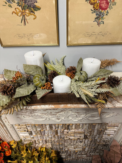 An autumn-themed fall centerpiece arranged on a rustic white fireplace mantel, featuring white pillar candles, green pumpkins, pinecones, dried ferns, and olive leaves, with framed botanical prints on the wall behind.