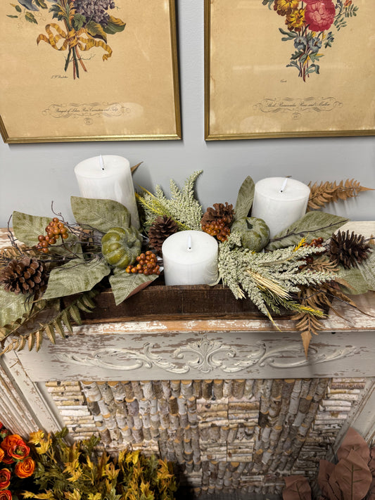 An autumn-themed fall centerpiece arranged on a rustic white fireplace mantel, featuring white pillar candles, green pumpkins, pinecones, dried ferns, and olive leaves, with framed botanical prints on the wall behind.