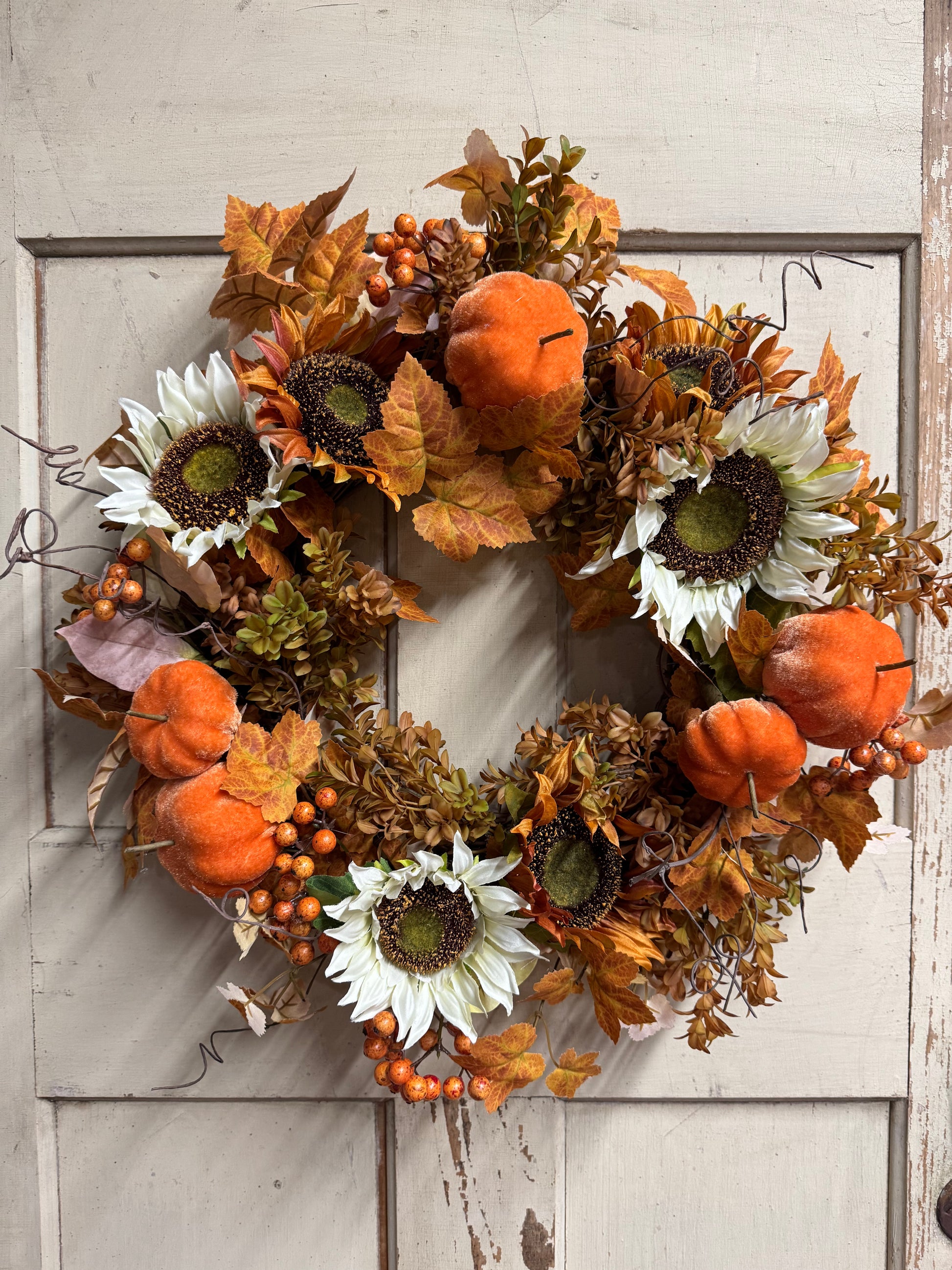 An autumn-themed artificial wreath hanging on a distressed white wooden door. The wreath features white sunflowers, orange pumpkins, brown and orange leaves, and small orange berries.