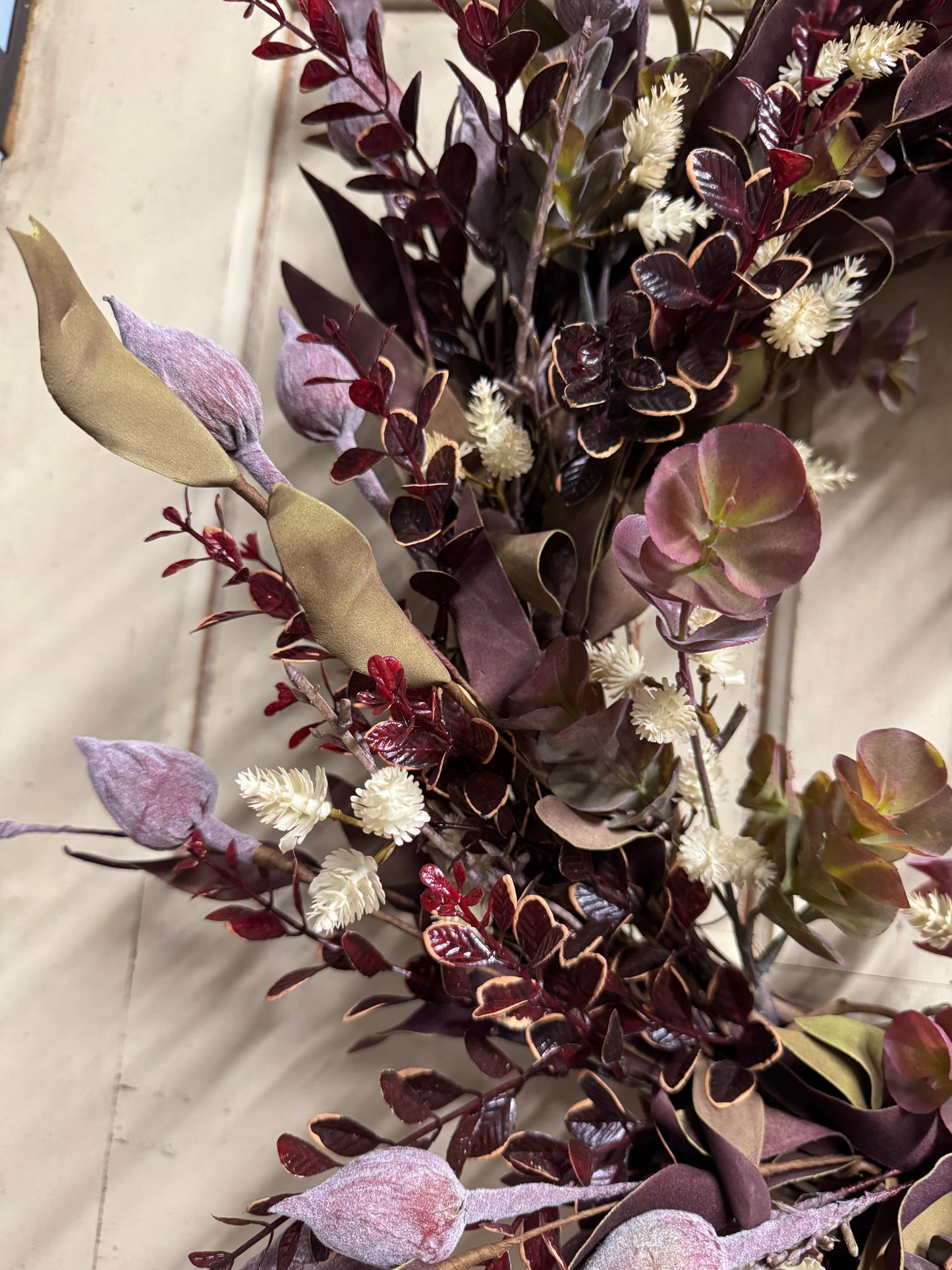 An artificial floral arrangement featuring dark burgundy and purple leaves, muted green leaves with gold veining, pale lavender buds, and small white fluffy flowers, photographed against a light beige background.