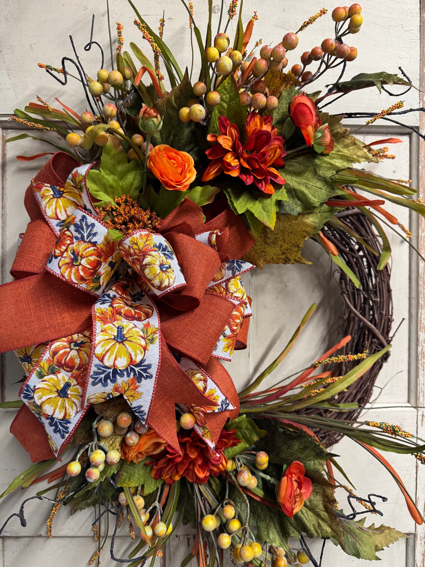 Handmade fall wreath with orange mums, burnt-orange and pumpkin-print bow, green grape leaves, autumn berries, and wispy grasses on a grapevine base