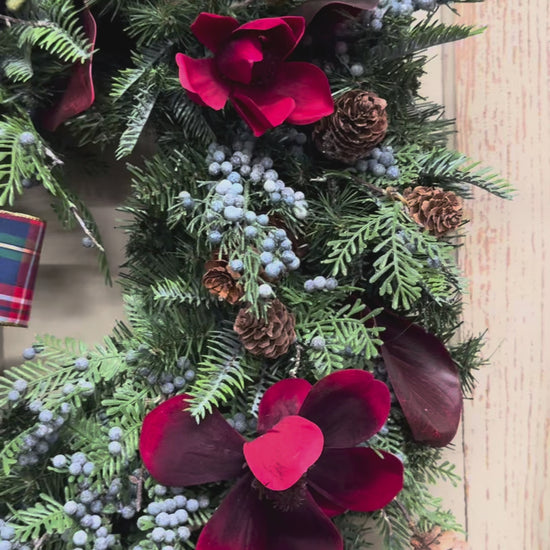 Video of Christmas pine wreath with red magnolias, burgundy leaves, blue juniper berries, pinecones, and a red, navy, and green plaid bow.”