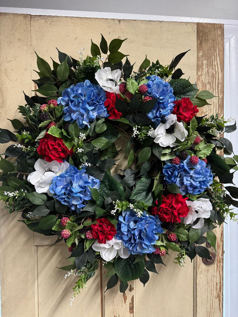 Handcrafted Raspberry Jubilee summer wreath with blue hydrangeas, red geraniums, white poppies, and raspberry stems on an 18-inch grapevine base
