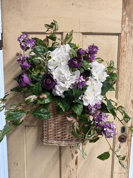 Floral arrangement with purple and white flowers in a woven basket against a wooden door.